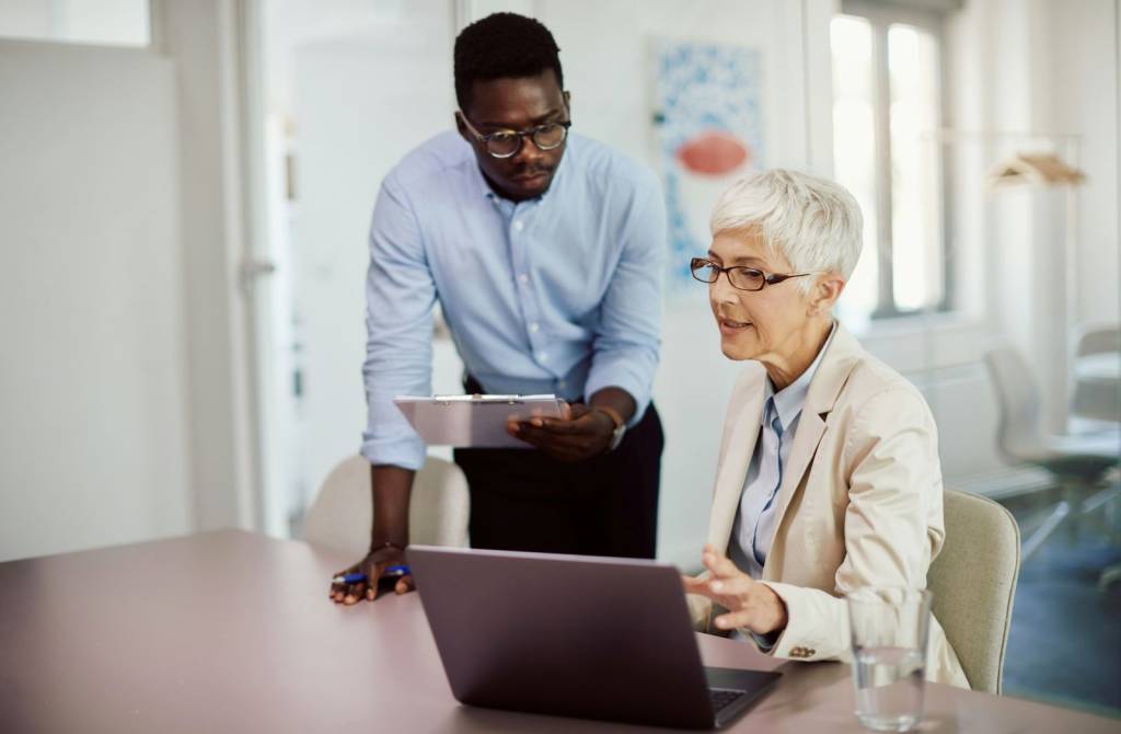 To personer på et kontor som har en samtale. En kvinne sitter ved et bord foran en laptop og viser noen på laptopen til en mann som står bak. Han holder et nettbrett. (Foto)