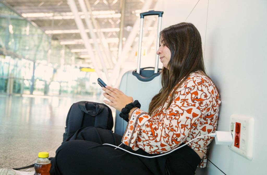Jente som sitter på gulvet på flyplass. Hun leser på mobilen. Mobilen er koblet til strøm via lader i stikkontakt på flyplassen. (Foto)