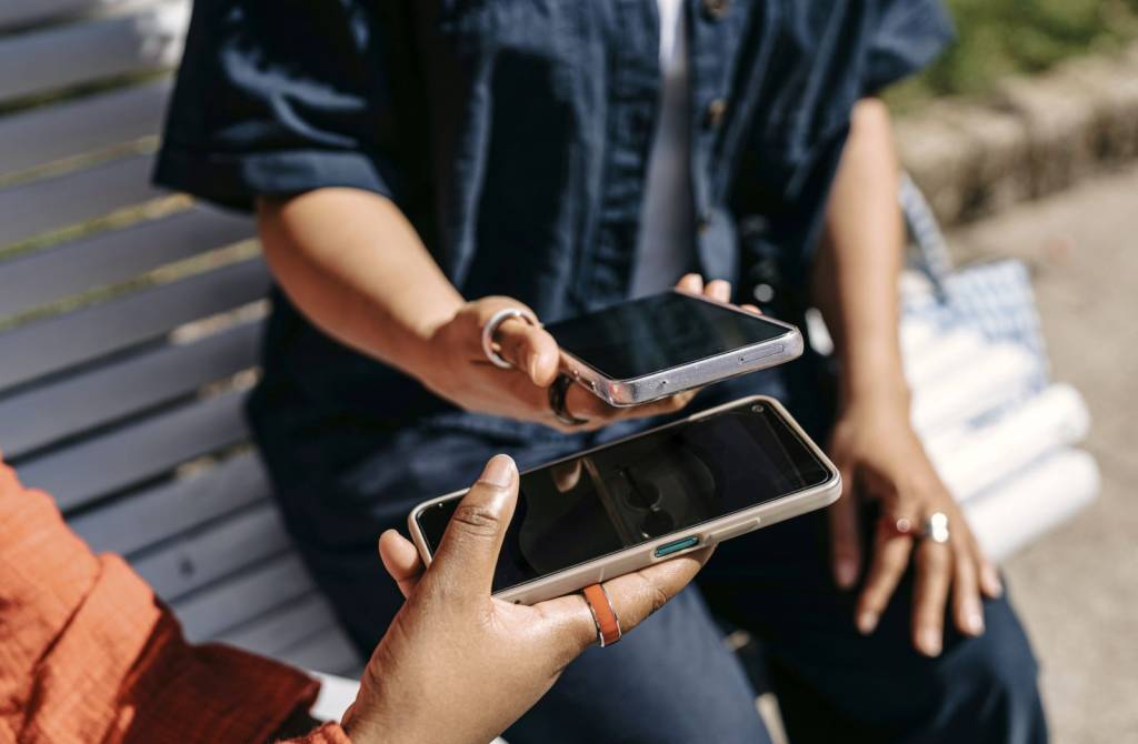 To personer med mobiltelefon sitter på en benk. Ansiktene vises ikke. I hendene har de hver sin mobil. De synkroniserer noe mellom de to mobilene. (Foto)