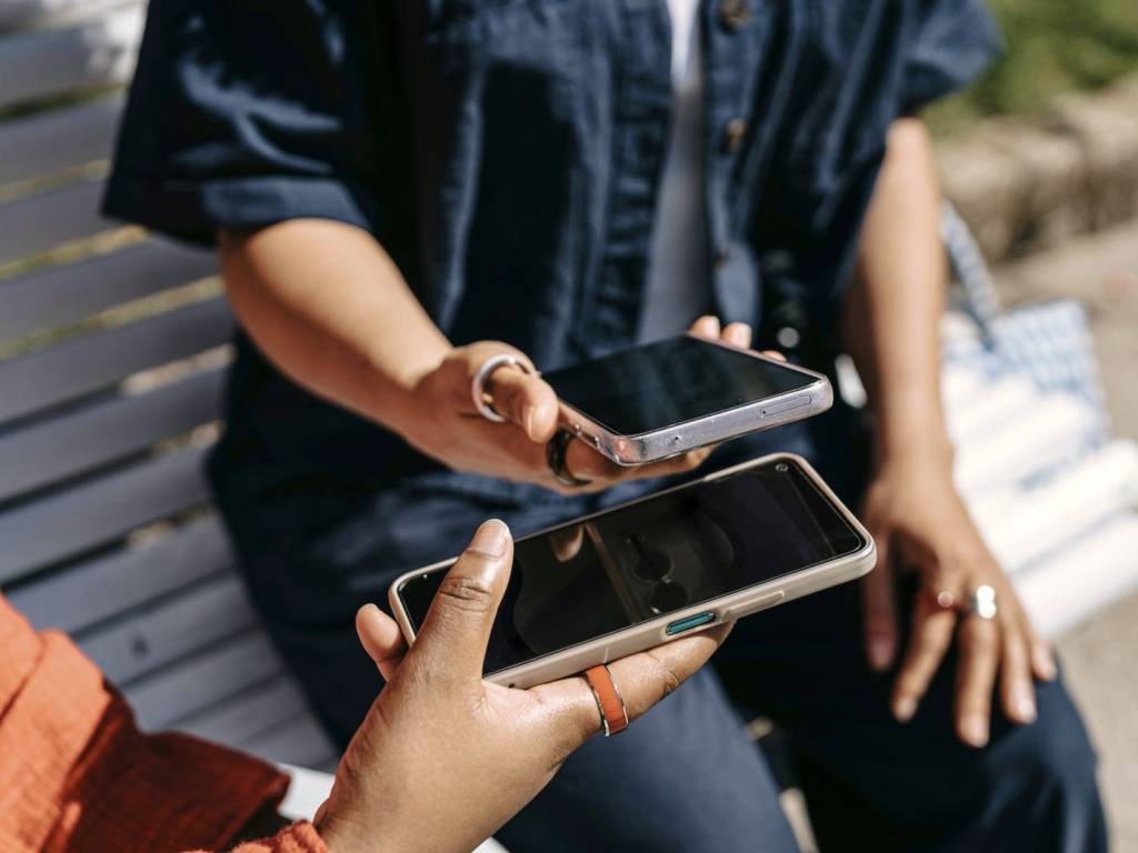 To personer med mobiltelefon sitter på en benk. Ansiktene vises ikke. I hendene har de hver sin mobil. De synkroniserer noe mellom de to mobilene. (Foto)